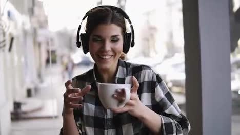 Joyful,-funny-young-girl-listening-to-the-music-in-headphones-and-dancing-with-a-cup-of-coffee-in-hand.-Blurred-street-on-the-background