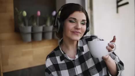 Brunette-girl-dancing-in-pleasure-with-a-cup-of-tea-and-headphones-listening-to-the-favourite-music.-Slow-motion.-Indoors