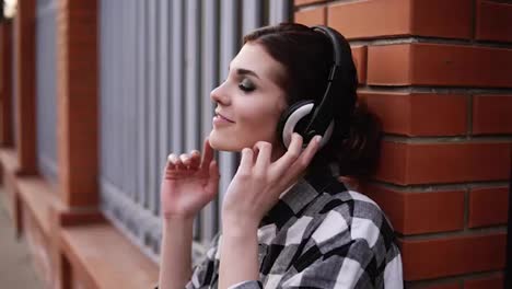 Beautiful-brunette-leaning-on-a-brick-fence-outdoors,-listening-to-music-on-headphones-with-eyes-closed.-Feels-the-music.-Emotionally-gesturing