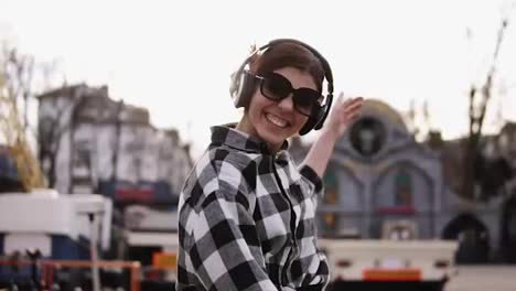 In-headphones-and-sunglasses,-trendy-girl-is-dancing-and-jumping-in-a-funny-way.-Smiling-and-laughing-with-open-mouth.-Outdoor.-Slow-motion