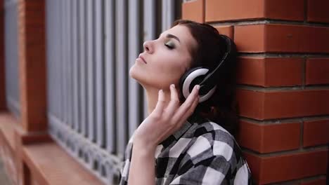 The-woman-pensively-listens-to-the-music-in-the-headphones,-leaning-on-the-wall-of-the-fence-in-the-street.-My-eyes-are-closed.-Make-up.-Close-up