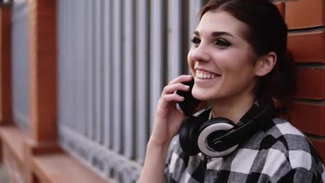 Extremely-close-footage.-Stunning,-young-girl-is-talking-on-mobile-phone-with-friend.-Standing-outside-leaning-on-a-brick-fence.-Trendy.-Happy.-Headphones-on-her-neck