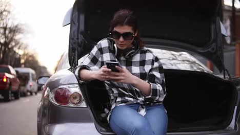 Woman-in-sunglasses-sits-in-open-trunk-of-car-in-jeans-and-plaid-shirt-along-the-street.-Typing-on-mobile-phone.-Headphones-on-her-neck.-Front-view
