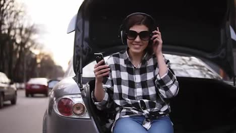 Smiling-brunette-listen-to-the-music-in-headphone-while-sitting-in-the-open-car's-trunk-among-the-road.-Cell-phone.-Good-mood