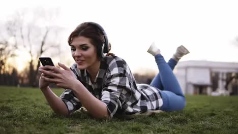 Happy-young-female-listening-music-on-smartphone-with-headphones-lying-on-green-grass-in-park.-Girl-having-fun-using-her-mobile-phone-outdoors