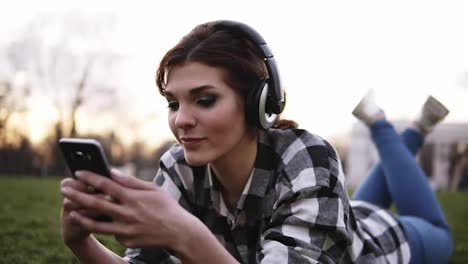 Young-girl-in-stylish-headphones-lying-on-a-grass,-listen-to-the-music-and-chatting-in-mobile.-Close-up