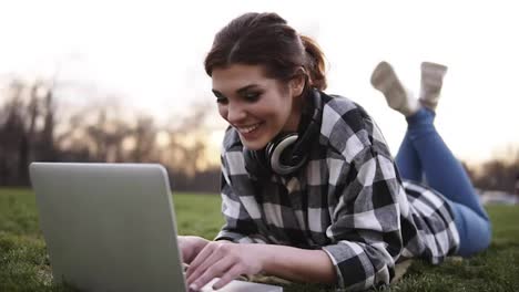 Beautiful-girl-lies-on-the-grass-in-the-park.-Uses-a-laptop,-chatting.-Headphones-on-the-neck.-She-has-a-good-time-in-communication.-Smiles,-happy.-Bright-day