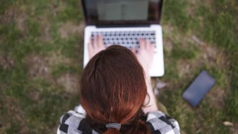 Aiming-footage-of-a-girl-with-a-hair-tail-sits-on-the-ground-in-a-park,-prints-on-a-blurred-laptop.-Mobile-is-on-the-grass-near.-Shooting-from-top-down
