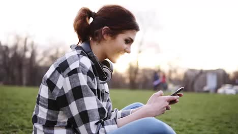 Girl-in-plaid-shirt-is-sitting-on-ground-in-park.-She's-messaging-with-a-mobile.-Happy,-smiling.-The-sun-is-shining-on-the-background.-Side-view