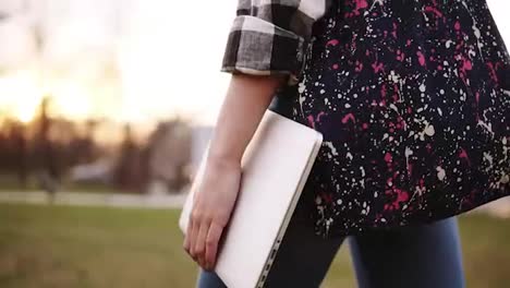 View-from-the-side-of-the-girl-in-jeans-walking-through-the-park-with-a-colored-bag-and-a-laptop-in-hand.-Close-up