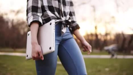 Slim-and-tender-girl-in-jeans-and-shirt-walking-by-green-side-outside,-holding-laptop-in-right-hand.-The-light-day-is-running-out.-Close-up.-Without-face