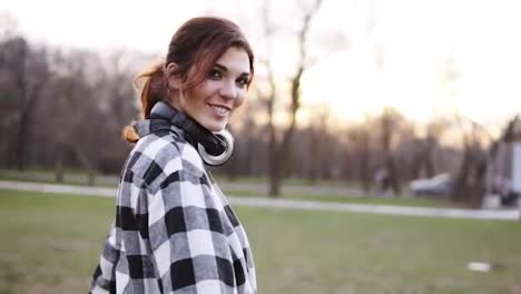A-beautiful,-self-confident-girl-with-headphones-walks-through-the-park-in-a-good-mood.-Spinning,-posing,-looking-at-the-camera.-Casual