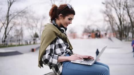A-young-trendy-brunette-is-sitting-on-a-bench-with-a-laptop-on-her-knees,-typing.-Headphones-on-the-neck,-glasses.-In-the-park-children-are-skating-on-scooters-and-bikes.-Side-view