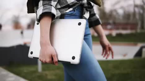 Close-up-of-an-elegant-girl-in-jeans-walks-through-the-park-with-a-laptop-in-her-hand.-Confident-walk.-Side-view.-No-face