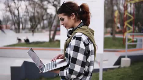 A-serene-beautiful-girl-walks-through-the-park.-With-one-hand-she-holds-a-laptop-and-messaging-with-someone.-Trendy-look.-Slow-motion