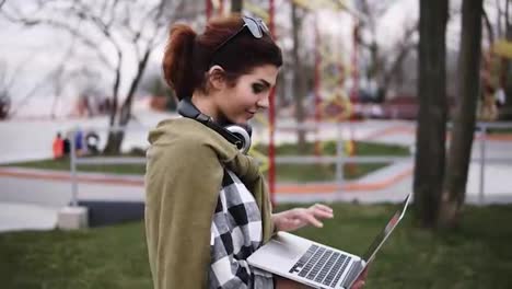 A-young-girl-with-sunglasses-on-her-head-and-a-plaid-shirt-walks-around-the-park,-her-laptop-is-in-her-hand.-She-goes-and-chatting-at-the-same-time,-typing-text.-Side-view