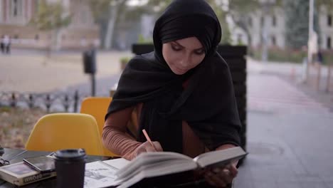 Una-Atractiva-Chica-Musulmana-Escribe-En-Un-Cuaderno-Sentada-En-Un-Café-Con-Un-Libro.-Estudiar-En-Un-Café-Al-Aire-Libre,-Tomar-Un-Descanso.-Universidad,-Autoeducación.-Vista-Frontal.-Camara-Lenta