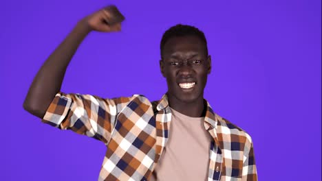 Strong-afro-american-young-man-wearing-plain-shirt-isolated-on-blue-background-in-studio-showing-muscules-at-camera,-sportsman.-People-sincere-emotions,-lifestyle-concept