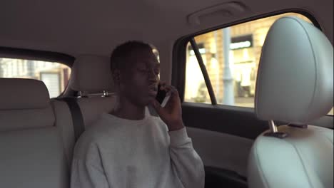 Calm-handsome-african-man-in-casual-clothes-talking-on-cellphone-while-riding-in-backseat-of-taxi-cab.-Cheerful-black-passenger-guy-answering-call-phone-during-road-trip-in-the-city