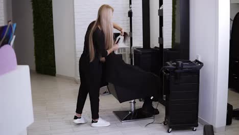 Full-length-of-blonde-woman-works-as-a-hairdresser-and-cuts-the-hair-ends-of-a-longhaired-female-customer-sitting-in-the-salon-chair.-Slow-motion.-Beauty,-hair-care-concept