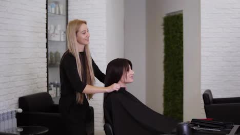 Female-hairdresser-has-just-finished-working-on-her-female-client-with-long,-straight-brown-hair.-Woman-is-smiling-and-glad-about-her-new-hair-style.-Hairdresser-removing-protective-cape.-Side-view