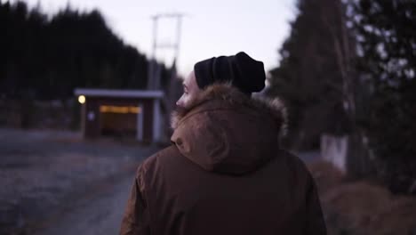 Footage-from-the-back-of-a-man-walking-down-the-countryside-road-on-cold-autumn-afternoon.-Wearing-black-hat-and-winter-coat-with-the-natural-fur-hood.-Evening-dusk.-Small-hut-on-the-background