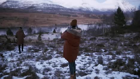Two-tourists-walking-in-slow-motion-through-the-countryside-towards-the-snowy-mountain-peaks-on-the-background.-A-person-on-a-winter-coat-trying-to-take-a-photo-of-beautiful-Norway-nature