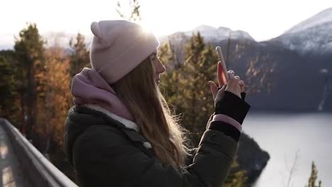 Woman-with-a-beige-hat-looking-on-horizon-from-the-road-and-taking-picture-of-the-incredible-beautiful-landscape-of-Norway-with-mountains-snowy-peaks-and-lake-using-her-smartphone.-Side-view