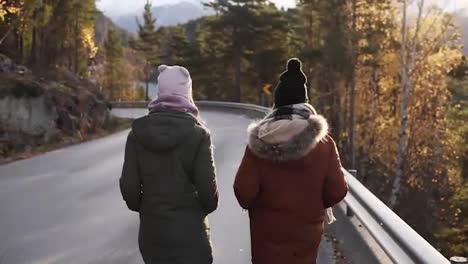 Two-female-tourists-walking-in-slow-motion-on-a-long-road-through-the-countryside-towards-the-snowy-mountain-peaks-and-golden-trees-on-the-background.-Rare-view