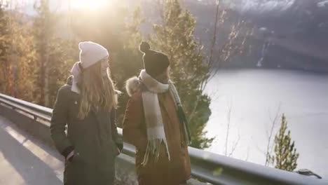 Zwei-Weibliche-Touristen-In-Wintermänteln-Laufen-In-Zeitlupe-Auf-Einer-Langen-Straße-Durch-Die-Landschaft-Zu-Den-Schneebedeckten-Berggipfeln,-Goldenen-Bäumen-Und-Dem-See-Im-Hintergrund.-Positive-Kaukasische-Mädchen-Erkunden-Die-Norwegische-Natur