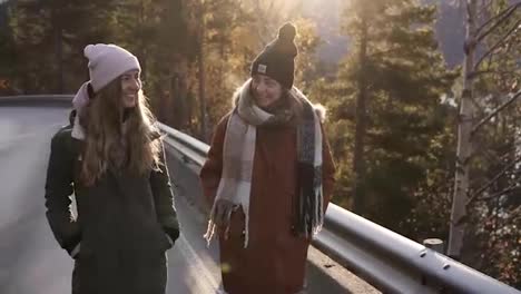 Positive-female-tourists-in-winter-coats-walking-in-slow-motion-on-a-long-road-through-the-countryside-towards-the-snowy-mountain-peaks,-golden-trees-and-lake-on-the-background.-Smiling-caucasian-girls-exploring-Norway-nature