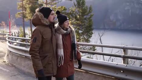 Tourists-in-winter-coats-and-black-hats-walking-in-slow-motion-on-a-long-road-through-the-countryside-towards-the-snowy-mountain-peaks-and-lake-on-the-background.-Smiling-caucasian-couple-exploring-Norway-nature