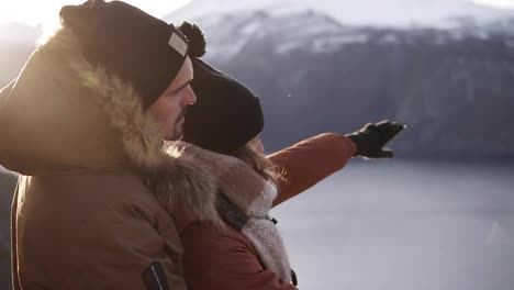 Young-couple-in-winter-coats-embracing,-people-hugging-looking-at-stunning-view-on-lake-and-mountains-peaks.-Admire-Beautiful-Mountain-Nature-Landscape.-Side-view