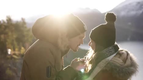 Young-couple-standing-embrace-kissing,-looking-landscape-sunny-winter-day.-Woman-and-man-in-warm-clothes-standing-in-front-beautiful-landscape-of-Norwegian-mountains-peaks-and-winter-lake.-Lens-flares-ona-background