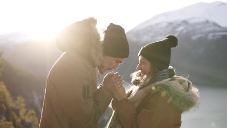 Young-couple-in-winter-coats-standingto-each-other,-holding-hands-towards-gorgeous-snowy-mountains-and-lake,-a-man-warming-his-girlfiends-hands-and-kissing.-Travel-concept.-Sun-strongly-shining-on-the-background
