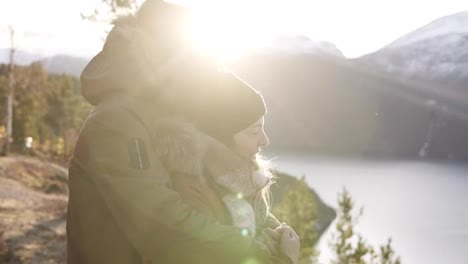 Happy-young-couple-in-winter-coats-embracing,-people-hugging-looking-at-stunning-view-on-lake-and-mountains-peaks.-Admire-Beautiful-Mountain-Nature-Landscape.-Lens-flares