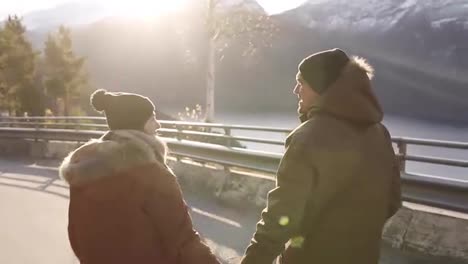 The-couple-looking-forward-to-the-adventure.-Together-they-run-along-an-asphalt-road-or-a-highway-in-winter-among-the-Norwegian-mountains.-Rare-view.-Sun-shining-and-lake-on-the-background