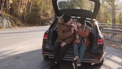 A-loving-couple-in-a-road-trip.-The-guy-and-the-girl-are-sitting-in-the-open-trunk-of-the-car,-eating-sandwiches-and-feeding-each-other.-Stop-outdoors-in-middle-of-the-road.-Cold,-sunny-autumn-day
