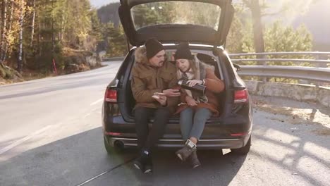 Loving,-young-couple-drinks-hot-tea-from-thermos-flask-sitting-in-car-trunk.-Happy-caucasian-couple-having-a-coffee-break-during-road-trip-in-countryside---drink-and-eating-sandwiches.-Admiring-the-nature