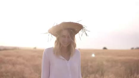 Attractive,-funny,-smiling-blonde-woman-in-white-shirt-and-straw-hat-posing-while-walking-by-wheat-field-on-sunny-summer-day