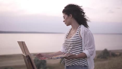 Side-view-of-an-attractive-curly-woman-artist-working-on-her-picture-with-easel-outdoors-near-the-lake.-Morning,-soft-wind-blowing-on-woman's-face