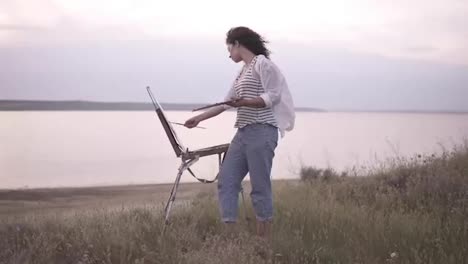 Full-length-footage-of-a-beautiful-girl-in-white-shirt-and-jeans-working-outdoors.-Paints-using-an-easel-and-oil-paints-on-the-meadow-in-front-the-lake.-Wind-blowing,-morning