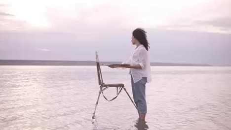 Gorgeous-view-of-a-young-curly-brunette-standing-in-the-water-with-easel-and-draing-her-the-surrounding-landscape.-Wearing-a-blue-jeans-and-white-shirt.-Full-length