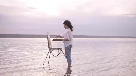 Accelerated-shooting,-a-girl-artist-in-the-water-till-ankles-draws-a-landscape-using-an-easel-and-a-palette.-Curly-girl-on-the-background-is-lake,-she's-in-casual-clothes:-jeans-and-a-white-shirt
