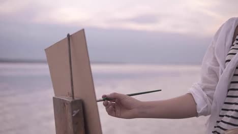 Close-up-of-an-artist's-art-process.-An-easel-and-palette.-Girl-is-putting-some-color-paints-on-canvas.-Blurred-lake-surround-on-the-background