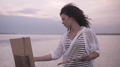 Close-up-of-a-gorgeous-woman-with-brunette-curly-hair-is-standing-in-front-the-lake-and-drawing-using-a-palette-and-an-easel.-Serious-and-concentrated.-Morning-clear-sky