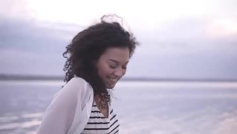 Beautiful-young-curly-brunette-walking-barefoot-in-sea-or-lake.-Happy,-smiling,-shy-female-wearing-strippes-t-shirt,-white-shirt-and-blue-jeans