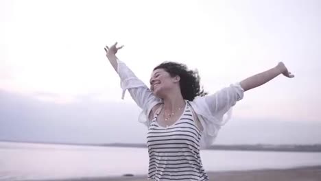 Portrait-of-a-gorgeous-curly-brunette-happily-running-near-the-sea-or-lake-with-outstretched-hands.-Wearing-white-casual-clothes.-Smiling,-happy,-freedom