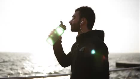 Young-attarctive-male-with-headphones-in-his-ears-drinking-water-from-plastic-fitness-bottle-after-workout.-Sport,-fitness,-youth-wellbeing.