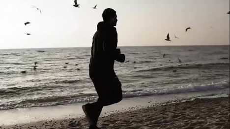 Slomotion-footage-of-a-sportive-male-running-along-the-sea-shore-with-seagulls-flying-on-the-background-view.